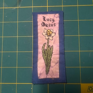 May include: A rectangular fabric patch featuring a light purple center panel with a dark blue border. The patch displays an embroidered white and yellow daffodil flower and the black embroidered text Lucy Dacus in a gothic-style font.