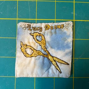 May include: A square fabric patch featuring a light blue and white tie-dye pattern. The patch displays an embroidered gold pair of scissors in the center and the text "Lucy Dacus" stitched in gold thread across the top portion of the fabric.