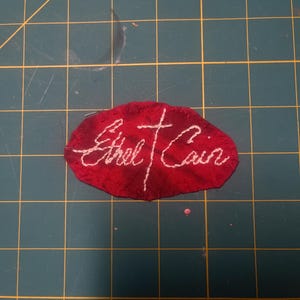 May include: An oval-shaped red fabric patch featuring white hand-stitched embroidery. The text reads Ethel Cain in a cursive script, with a vertical cross separating the two words. The edges of the red fabric are slightly frayed and uneven.