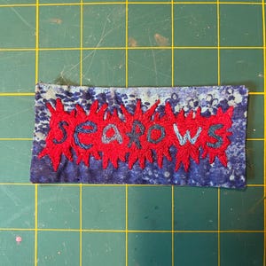 Könnte beinhalten: Ein rechteckiger Stoffaufnäher mit einem blau-weiß gesprenkelten Hintergrund. In der Mitte befindet sich ein gezacktes, sternförmiges rotes Stickdesign, bei dem das Wort "seapows" mit dunkelblauem Garn in die Mitte gestickt ist.