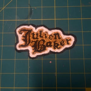 May include: A fabric patch featuring the text Julien Baker embroidered in black thread. The lettering is surrounded by a textured gold-colored fill, a white border, and an outer black edge, creating a distinct layered appearance for the design.