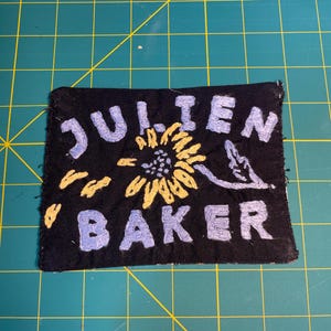 May include: Black embroidered patch with the words "JULIEN BAKER" in light blue, with a yellow sunflower in the center. The patch is rectangular and appears to be handmade.
