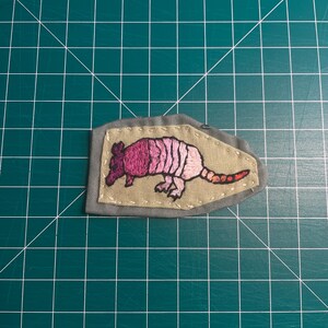 May include: A pink and orange embroidered armadillo patch on a gray fabric background.