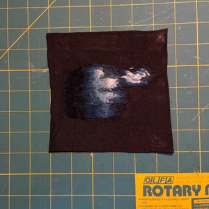 May include: A square black fabric patch featuring an embroidered design of a dark, swirling cloud-like shape in shades of deep blue, navy, and white, with a smaller, lighter puff of white and blue thread extending to the right side.