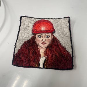 May include: Embroidered portrait of a person wearing a red hard hat and with long, red hair. The artwork is framed with a black border and set against a light gray background. The embroidery captures facial features and hair texture.