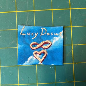 May include: A square fabric patch featuring a blue sky with white clouds print. The text "Lucy Dacus" is embroidered in light tan thread at the top. Below the text, an infinity symbol and a heart are embroidered in a dark red thread.