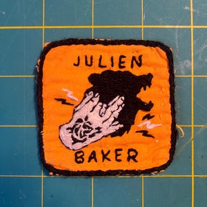 May include: A square embroidered patch with an orange background and a black border. It features a black silhouette of a bear and a white hand making a gesture. The text JULIEN is embroidered in black above the bear, and BAKER is below.