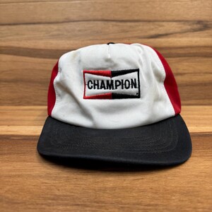 May include: A white, red, and black baseball cap with a black embroidered logo that says "CHAMPION" on the front.