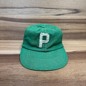 May include: A vintage green baseball cap with a white "P" on the front. The cap has a curved brim and is made of a textured fabric. The hat is likely from the early to mid-20th century.
