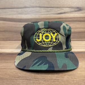 May include: A camouflage baseball cap with a yellow embroidered globe and the word "JOY" inside the globe.