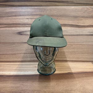 May include: Olive green baseball cap with a curved brim. The cap is displayed on a clear glass head-shaped form against a wooden background. The cap has a structured crown and a slightly faded appearance.