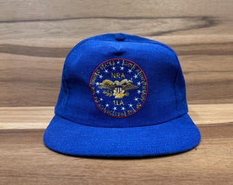 May include: A blue corduroy baseball cap with an embroidered patch featuring the NRA logo, an eagle with a shield, and the text "United States - 150th Anniversary - The Freedom to Keep and Bear Arms - NRA".