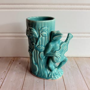 May include: A teal ceramic mug featuring a sculpted frog playing a banjo. The mug has a textured surface resembling a tree trunk with leaves and branches. The frog is depicted in a seated position, holding the instrument against its body.