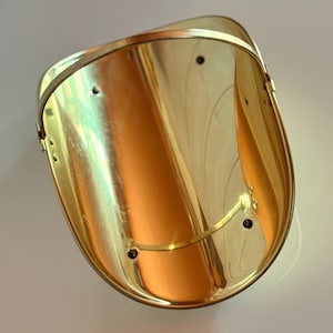 May include: A polished brass scoop with a curved, open-top design and a rounded handle. The interior surface is smooth and reflective, featuring four small circular holes arranged symmetrically along the base and sides of the metal structure.