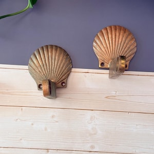 May include: Two metallic wall hooks shaped like scallop shells with a weathered bronze finish. Each hook features a fan-like ribbed design with a protruding curved arm at the base and two small circular screw holes for mounting on a flat surface.