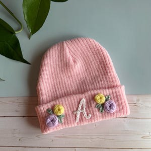 May include: A pink ribbed knit beanie with embroidered floral accents and the letter "A". The flowers are yellow and purple. The hat is displayed on a light wood surface with a green leafy plant in the background.