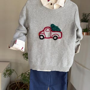 May include: A gray sweater with a red truck and green tree design, paired with a collared shirt and navy blue shorts. The sweater has long sleeves with white cuffs. The outfit is displayed on a hanger.