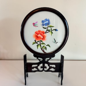 May include: A round, embroidered floral artwork featuring a red peony, blue flower, and butterflies. The artwork is set in a dark wood frame and displayed on a matching stand. The embroidery is detailed and colorful.