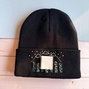May include: Black beanie with embroidered mountain and tree design. The design includes white mountains, white dots, and green trees. A blank square is in the center of the design. The beanie is made of a soft, knitted material.
