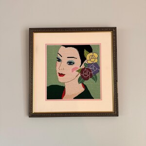 May include: Framed needlepoint art featuring a woman's portrait with black hair, red lips, and blue eyeshadow. The artwork includes flowers in her hair and is set against a green background, framed in a gold-toned frame.