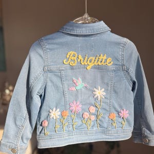 May include: Light blue denim jacket with embroidered flowers and a hummingbird design. The name "Brigitte" is embroidered in yellow thread across the back. Features a classic collar and button closures.