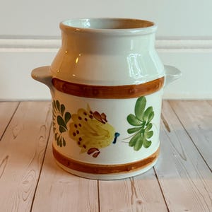 May include: A white ceramic jar with two side handles, featuring a hand-painted design of yellow and green floral motifs. The jar is decorated with two horizontal brown bands encircling the body, providing a contrast to the white base.