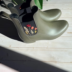 May include: Olive green rain boots with a low ankle cut. The boots feature an embroidered floral design in red, yellow, and white on a black elastic panel. The boots are made of a waterproof material and are suitable for wet weather.