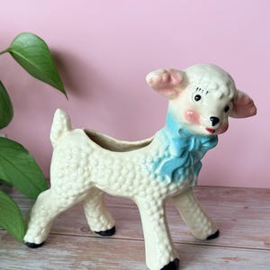May include: A vintage ceramic lamb planter in cream with a blue scarf and black hooves. The lamb has pink ears and cheeks, and a small opening on its back for plants. The planter is on a wooden surface.