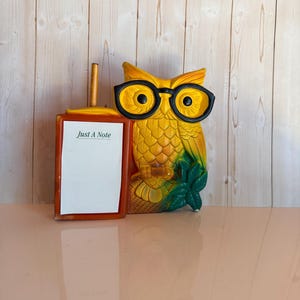 May include: A yellow owl figurine wearing black glasses, featuring a rectangular orange frame attached to its side. The frame holds a white notepad with the text "Just A Note" printed in green. A yellow pencil is inserted into the top of the frame.