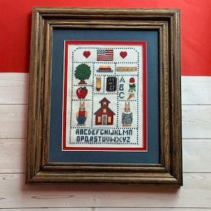 May include: A framed cross-stitch sampler featuring a schoolhouse, two rabbits, a tree, an apple, a pencil, a ruler, books, and the American flag. The design includes the alphabet in black letters and two red hearts, all set within a dark wood frame.