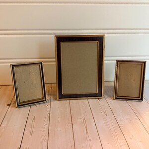 May include: Three rectangular picture frames with dark brown wood-grain centers and thin gold-toned metallic borders. The frames are arranged in a row, with one larger frame positioned in the center flanked by two smaller frames of identical size.