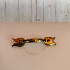May include: A polished brass drawer pull featuring a central horizontal bar with a smooth finish and two flared, tulip-shaped ends. Each end is accented with a small, raised square pyramid detail centered within the metal petal design.