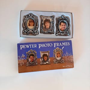 May include: A set of three silver-toned pewter photo frames with intricate floral detailing. The frames are presented in a white box and include a blue cardboard packaging sleeve labeled PEWTER PHOTO FRAMES Set of Three with images of the frames.