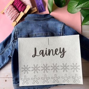 May include: A denim jacket with a white canvas patch that reads "Lainey" in black script, with star-shaped designs below. Pink and purple yarn spools and a needle are also visible.