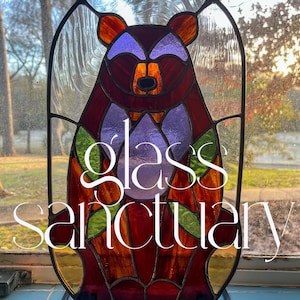 Pode incluir: Uma janela de vitral representando um urso com detalhes em roxo, vermelho e verde. A janela é enquadrada em metal preto e tem as palavras "santuario de vidro" escritas nela.