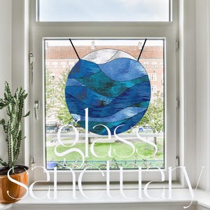 Puede incluir: Una ventana de vidrio emplomado colgada en un marco de ventana blanco. El vidrio emplomado es un círculo con olas azules y verde azulado. El texto "glass sanctuary" está escrito en letras blancas debajo de la ventana.