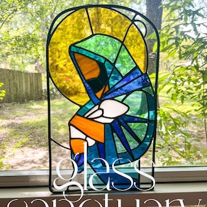 Może przedstawiać: Panel witrażowy przedstawiający postać religijną ze złotą aureolą oraz niebieskimi, zielonymi i pomarańczowymi szatami. Dzieło oprawione jest w czarną metalową ramę, a u dołu widnieje napis "Glass Sanctuary".