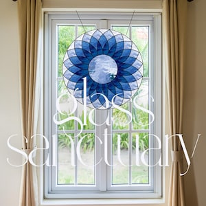 May include: A stained glass window with a blue and white mandala design. The window is framed in white and is set in a room with beige curtains.