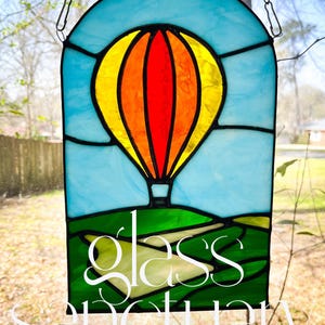 May include: Stained glass panel featuring a colorful hot air balloon against a blue sky. The balloon is yellow, orange, and red. The panel includes green rolling hills and the text "Glass Sanctuary".