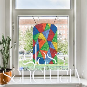 May include: A colorful stained glass window hanging in a window frame. The glass is made up of many small pieces of glass in different colors, including red, orange, yellow, green, blue, and purple. The window is hanging in a white window frame with a white wall behind it. The text "glass sanctuary" is written in white letters on the bottom of the image.