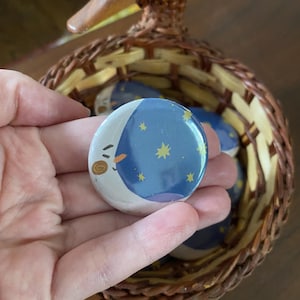 May include: A round button with a blue night sky design featuring yellow stars and a smiling crescent moon. The button is held in a hand, with more buttons visible in a woven basket with a duck-shaped handle.