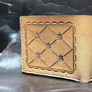 Op de afbeelding: Een bruine leren portemonnee met een decoratief patroon van ruitvormige figuren en bladeren. De portemonnee is gesloten en heeft een vouwdesign.