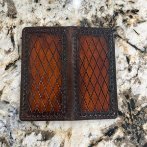 May include: A rectangular brown leather wallet featuring two distinct panels, each embossed with a diamond grid pattern and bordered by a decorative scalloped edge. The leather displays a gradient of dark brown edges transitioning to a lighter tan centre.