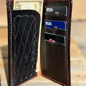 Peut inclure: Portefeuille en cuir noir avec coutures rouges. Le portefeuille est ouvert, révélant un porte-billets avec un motif décoratif et plusieurs fentes pour cartes contenant des cartes de crédit. Un billet de dix dollars américains est partiellement visible.