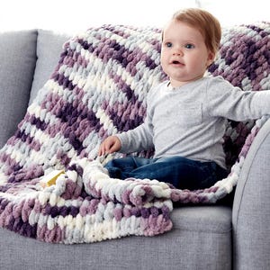 Peut inclure: Un bébé assis sur un canapé gris avec une couverture en tricot moelleuse et épaisse dans des tons de violet, blanc et gris. La couverture est faite de fil épais et a un aspect texturé et moelleux.