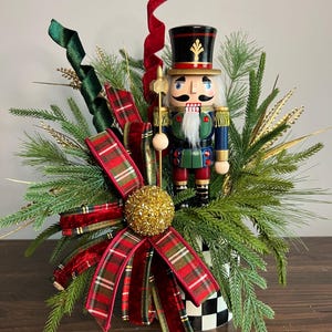 May include: A decorative nutcracker figure wearing a black hat and blue uniform, holding a gold staff. It is surrounded by green pine branches, red and green plaid ribbon, a gold glittered sphere, and a red velvet ribbon spiral.