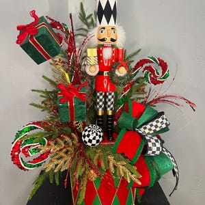 May include: A decorative nutcracker centerpiece featuring a red and green drum base, evergreen branches, and gift boxes. The nutcracker wears a black and white hat, red jacket, and checkered pants, surrounded by red and green ribbons and ornaments.