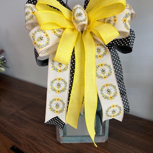 Könnte beinhalten: Eine dekorative Schleife mit gelben, schwarzen und cremefarbenen Bändern. Die Schleife hat ein zentrales gelbes Band und ist mit Bienen- und Sonnenblumenmustern verziert. Schwarze Polka-Punkte-Bänder sorgen für Kontrast. Für die Heimdekoration.