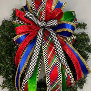 Puede incluir: Un lazo decorativo de varias capas con una mezcla de cintas en rojo, verde, azul y purpurina dorada, acentuado con un patrón de cuadros blanco y negro y un diseño de tartán en rojo, verde y azul, sujeto a una corona de pino artificial.