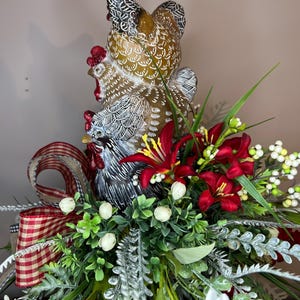 May include: A decorative centerpiece featuring three ceramic chickens in varying sizes, with brown, white, and black detailing. The arrangement includes red lilies, white berries, and greenery, accented with a red and white checkered ribbon.
